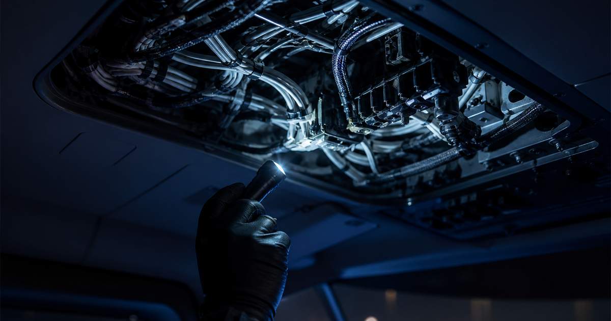 Overhead panel de cockpit d'hélicoptère ouvert sur son faisceau de câbles électriques, lampe d'inspection tenue par une main gantée